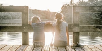 Two children sitting side by side sitting on a dock looking out onto the water