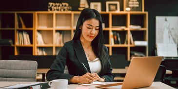 woman in an office writing on notepad with laptop